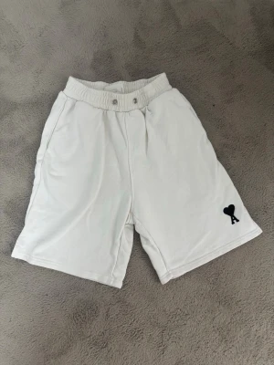Vita shorts med hjärtdetalj - Säljer ett par vita shorts med bred resårmidja och två knappar framtill. På vänster ben finns en svart broderad detalj med ett hjärta och bokstaven A. Shortsen har avslappnad passform och sidofickor. Perfekta för sommardagar.