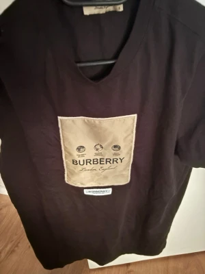 Svart Burberry t-shirt med beige patch - Säljer en svart t-shirt från Burberry med en stor beige patch på bröstet där Burberry-loggan och texten 'London, England' syns. T-shirten har rund hals och korta ärmar. Perfekt för dig som gillar stilrena designer med en twist.