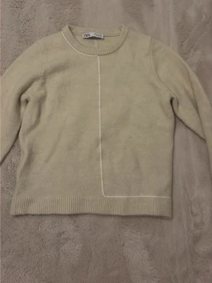 Beige stickad tröja från ZARA - Säljer en beige stickad tröja från ZARA med rund halsringning och en unik vit linje som går vertikalt och horisontellt framtill. Tröjan har långa ärmar och ribbade muddar vid ärmslut och nederkant. Perfekt för en stilren och enkel look.