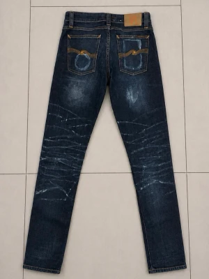 Nudie Jeans 28/32 - Säljer ett par mörkblå nudie jeans med coola slitningar och whiskers på framsidan och baksidan. Jeansen har raka ben, klassiska fem fickor och kontrastsömmar. Materialet är denim i bomull och passformen är smal. Perfekta för en avslappnad och trendig look.