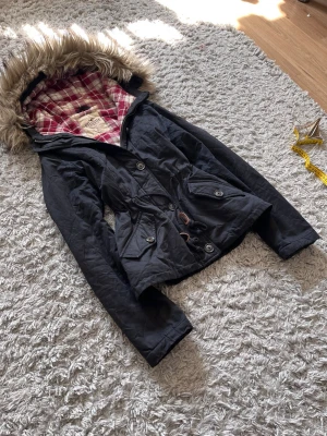 Svart parkas med fuskpäls och rutigt foder - Snygg svart parkas med huva och fuskpäls, dragsko i midjan och stora fickor framtill. Insidan har ett vadderat rödvitt rutigt foder som ger extra värme och stil. Perfekt för kalla dagar och riktigt mysig att bära.
