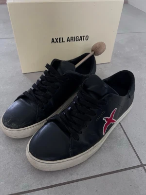 Svarta Axel Arigato sneakers med röd fågel - Snygga svarta sneakers från Axel Arigato i läder med vit sula och svart snörning. På sidan finns en broderad röd fågel och diskret Arigato-tryck. Klassisk låg modell som passar till det mesta och ger en clean look.