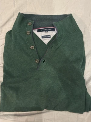 Grön tröja från Tommy Hilfiger - Mörkgrön långärmad tröja från Tommy Hilfiger med ribbade muddar och knappslå vid halsen. Tröjan har en klassisk passform och är tillverkad i ett mjukt material som känns skönt mot huden. Perfekt för dig som gillar stilrena och bekväma plagg. Aldrig använd