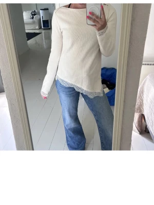 Vit/beige långärmad topp med spetsdetaljer - Säljer en vit långärmad topp med ribbad struktur och fina spetsdetaljer vid ärmslut och nederkant. Toppen har en rak passform och liten slits i sidan, vilket ger en snygg och avslappnad look. Perfekt att styla med jeans eller kjol.