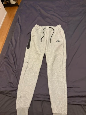 Gråa mjukisbyxor från Nike - Snygga ljusgrå mjukisbyxor från Nike med svarta detaljer och logga på benet. Byxorna har dragsko i midjan, muddar vid bensluten och praktiska fickor. Perfekta för chill eller träning, riktigt sköna och stilrena.