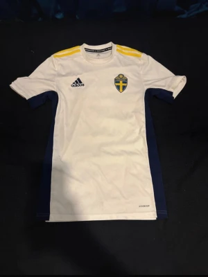 Svensk landslagströja Adidas vit - Säljer en vit svensk landslagströja från Adidas med blå detaljer på sidorna och gula ränder på axlarna. Tröjan har korta ärmar, rund hals och Sveriges landslagsemblem på bröstet. Perfekt för fotbollsträning eller att bära på matchdag.