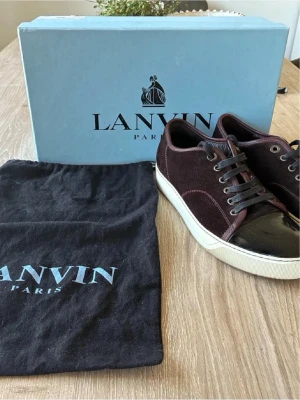 Vinröda Lanvin  - Snygga vinröda Lanvin i  mocka med svart lackad tå och vita sulor. Skorna har svarta skosnören och metallfärgade öljetter. Klassisk låg modell som passar till många olika stilar. Kommer med originalkartong och dustbag.