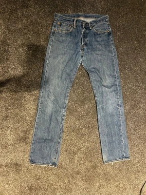 Levis 501 - Ett par klassiska blå jeans med rak passform och hög midja. Jeansen har fem fickor, gylf med dragkedja och knapp samt rå, avklippt kant vid bensluten för en trendig look. Materialet är kraftig denim i 100% bomull.