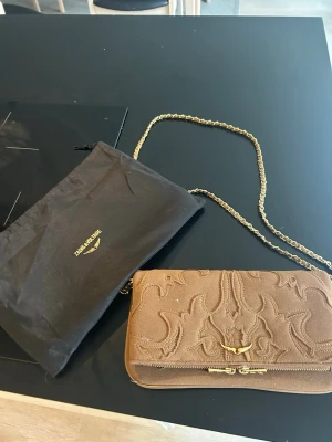 Brun kuvertväska med kedjerem - Snygg brun kuvertväska från Zadig & Voltaire med dekorativa broderier på framsidan och guldfärgade detaljer. Väskan har en elegant kedjerem i guld och ett litet emblem framtill. Perfekt för dig som vill ha en unik accessoar.