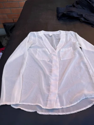Vit transparent blus från Vila - Snygg vit transparent blus från Vila i storlek XS. Blusen har lång ärm, två bröstfickor och en avslappnad passform. Tillverkad i ett tunt, lätt material som ger en luftig känsla. Perfekt att styla lager på lager eller med ett linne under.