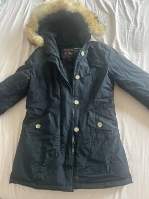 ÄKTA Woolrich jacka med rävpäls - Snygg svart parkas från Woolrich med huva och fuskpälsdetalj. Jackan har dragkedja och knappar framtill samt två fickor med lock. Perfekt för kalla dagar och har en klassisk, tidlös look.