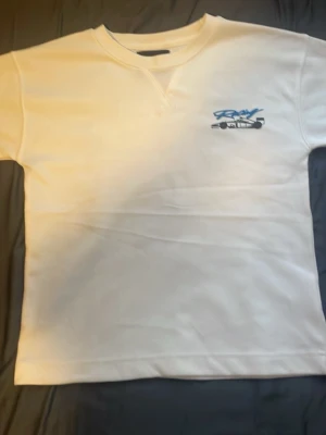 Vit t-shirt med racingtryck från peoples market  - Original priset är 400kr Säljer en vit t-shirt med blått och svart racingtryck fram och stort motiv av en Formel 1-bil på ryggen. T-shirten har rund hals och korta ärmar. Perfekt för dig som gillar motorsport och vill ha en clean men sportig look. 