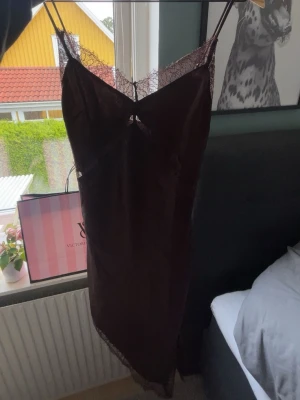 Brun silkesklänning med spets - Brun klänning av silke med tunna axelband och spetsdetaljer vid urringning och nederkant. Klänningen har en slim passform och är gjord i ett glansigt satinliknande material för en elegant look.