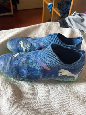 Blåa fotbollsskor från Puma - Säljer ett par blåa fotbollsskor från Puma med stickad strumpliknande öppning och vita detaljer. Skorna har en modern design med stora vita Puma-loggor och mönstrade sidor. Sulan är ljusgrön och ovandelen är i ett ventilerande syntetmaterial.