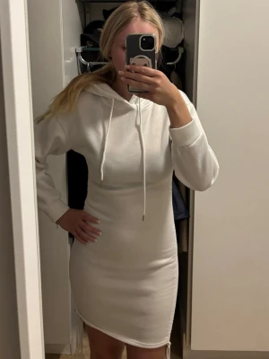 Vit hoodieklänning med snörning - Säljer en vit hoodieklänning med långa ärmar och huva. Klänningen har snörning i halsen och är i mjukt material, perfekt för en avslappnad stil. Modellen är kort och har en slim passform som sitter snyggt på kroppen.