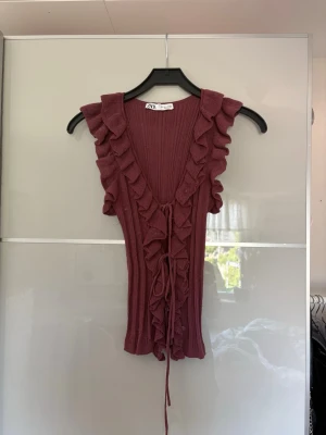 Vinröd ribbad topp med volang - Populär vinröd ribbad topp med volangdetaljer längs ärmhålen och halsringningen. Toppen är ärmlös, har djup v-ringning och snörning framtill. Perfekt för dig som vill ha en söt och trendig look.