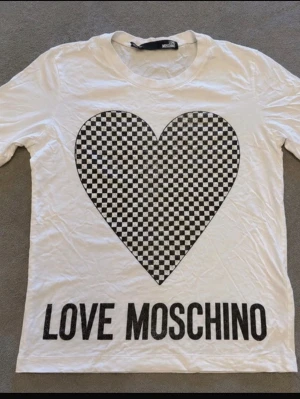 Vit Love Moschino t-shirt med hjärta - Snygg vit t-shirt från Love Moschino med ett stort schackrutigt hjärta i svart och vitt på bröstet. T-shirten har rund hals och korta ärmar. Materialet känns mjukt och skönt, troligen bomull. Perfekt för dig som gillar statement-plagg.