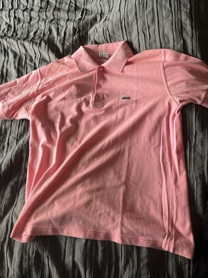 Rosa pikétröja från Lacoste - Säljer en klassisk rosa pikétröja från Lacoste med korta ärmar och krage. Tröjan har knappar framtill och den ikoniska krokodilloggan på bröstet. Perfekt för dig som gillar en sportig och stilren look. En riktigt snygg sommartröja!