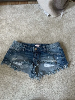Blå slitna jeansshorts med fransar - Säljer ett par blå jeansshorts med slitna detaljer och fransig kant. Shortsen har klassisk femficksmodell, bälteshällor och knappstängning. Perfekta för varma dagar och ger en avslappnad look.