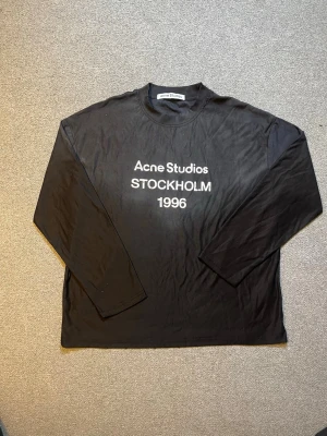 Svart långärmad t-shirt Acne Studios - Svart långärmad t-shirt från Acne Studios med vit text 'Acne Studios STOCKHOLM 1996' på bröstet. Klassisk passform och rund halsringning. Perfekt för dig som gillar stilrena och tidlösa plagg.