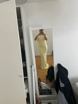 Ljusgul lång offshoulder klänning - Elegant ljusgul långklänning med offshoulder-design och figurnära passform. Klänningen har en asymmetrisk axel och mjukt draperad överdel som ger en exklusiv känsla. Perfekt för speciella tillfällen när du vill sticka ut.