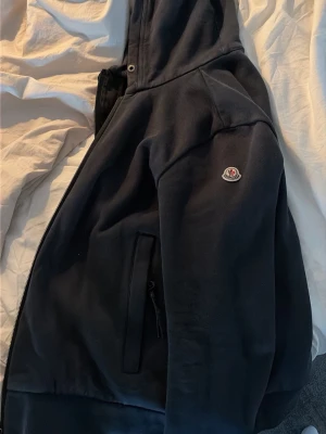 Svart hoodie från Moncler med huva - Säljer en svart hoodie från Moncler med dragkedja, huva och fickor på sidorna. Klassisk Moncler-logga på ärmen och mjukt material på insidan. Perfekt för dig som vill ha en stilren och exklusiv hoodie till vardagslooken. Kan dock vara lite för liten för dig som har storlek M, jag gillar den men nu är det daxs för den att få en ny ägare