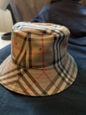 Burberry bucket hat med rutigt mönster - Snygg bucket hat från Burberry med klassiskt beige rutigt mönster i svart, vitt och rött. Hatten har rund form och är tillverkad i ett slätt tygmaterial. Perfekt accessoar för att lyfta din stil.
