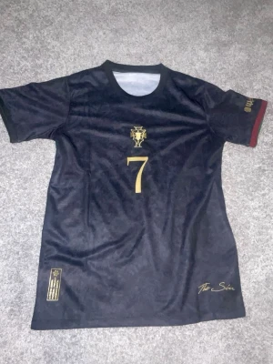 CR7 t-shirt- ”THE SIU” - Pris kan diskuteras! Snygg svart fotbollströja med nummer 7 i guld på både fram- och baksidan. Tröjan har guldfärgade detaljer, emblem på bröstet och texten 'The Siu' nedtill. Ärmarna har diskreta gröna och röda ränder. Perfekt för dig som gillar sportig stil.