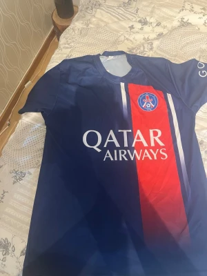 PSG matchtröja blå/röd - Snygg PSG matchtröja i blått med röd vertikal rand och vita detaljer. Tröjan har klubbmärke på bröstet och Qatar Airways tryckt på framsidan. Perfekt för fotbollsträning eller att bära till vardags för att visa ditt stöd för laget.