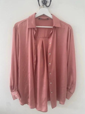  långärmad skjorta i satinlook - Säljer en snygg rosa skjorta med lång ärm och klassisk krage. Skjortan har en slät satinliknande yta och knappar framtill. Passar perfekt för dig som gillar en stilren och trendig look.
