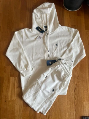 Vit hoodie och byxa loungewear-set - Snyggt vitt loungewear-set med hoodie och matchande byxor. Hoodien har dragkedja, snörning och fickor framtill samt en liten broderad logga på bröstet. Byxorna har resår i midjan och samma broderade logga. Perfekt för chill dagar hemma.