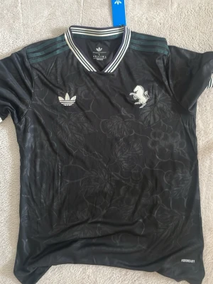 Svart Adidas topp med blommönster - Svart topp från Adidas med diskret blommönster och vita detaljer. Klassisk krage med ränder och korta ärmar. Adidas-logga och lejonmärke framtill. Perfekt för en sportig och trendig look.