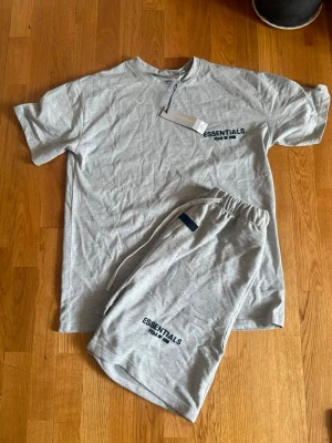 Grått Essentials Fear of God set - Säljer ett grått set från Essentials Fear of God med t-shirt och shorts. Båda plaggen har svart logotyptryck framtill och t-shirten har även stor logga på ryggen. Tillverkade i mjukt bomullsmaterial, perfekt för chill eller loungewear.