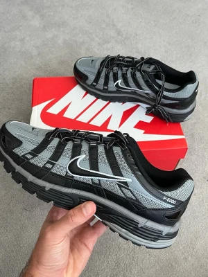Nike P-6000 svart/grå sneakers - Nike P-6000 sneakers i svart och grått med vit swoosh. Skorna har en sportig design med mesh och syntetiska överdrag, samt robust sula för extra komfort. Perfekta för dig som gillar streetwear och vill ha en retroinspirerad look.