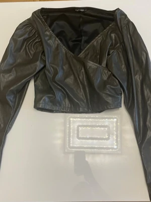 Svart omlottopp i skinnimitation - Snygg svart omlottopp från Gods Collection i skinnimitation. Toppen har långa puffärmar och croppad passform med v-ringad hals. Perfekt för dig som vill ha en edgy och trendig look.