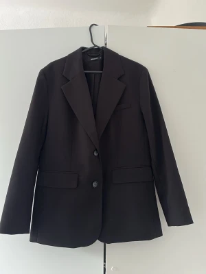 Svart klassisk kavaj med två knappar - Stilren svart kavaj med två knappar framtill och klassiska slag. Kavajen har två fickor med lock och en bröstficka. Perfekt för en dressad look och passar till många olika outfits. Materialet känns slätt och har en snygg passform.