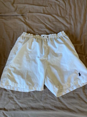 Badshorts från Polo Ralph Lauren - Klassiska vita shorts från Polo Ralph Lauren med elastisk midja och broderad logga på benet. De har en avslappnad passform och är tillverkade i bomull, perfekta för varma dagar.