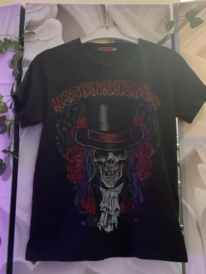 Svart t-shirt med döskalle och rosor - Cool svart t-shirt med tryck av en döskalle i hög hatt, omgiven av röda rosor och texten 'Maestro' i gotisk stil. T-shirten har korta ärmar och rund hals. Perfekt för dig som gillar rockig och edgy stil.