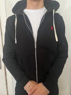 Svart zip hoodie med vit dragsko - Snygg svart hoodie med dragkedja från Ralph Lauren. Har vit dragsko i huvan och röd logga broderad på bröstet. Tillverkad i mjukt material och har två fickor framtill. Perfekt för en avslappnad och stilren look.Priset går att diskutera vid snabb affär, finn de frågor så är de bara o skriv 😊