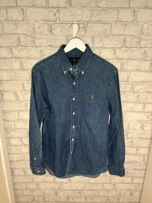 Ralph Lauren skjorta - Ralph Lauren jeansskjorta i klassisk mellanblå tvätt med broderad logga och slim fit-passform. Tillverkad i mjuk 100 % bomull som ger en bekväm känsla och passar lika bra uppknäppt över en t-shirt som för sig själv. Mycket fint skick. Hör gärna av dig vid intresse! 🙌
