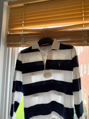 Randig rugbytröja från Polo Ralph Lauren - Säljer en långärmad rugbytröja från Polo Ralph Lauren med breda marinblå och vita ränder. Tröjan har klassisk krage med tunna ränder på insidan och den ikoniska broderade loggan på bröstet. Perfekt för en sportig och avslappnad stil. Står M men den sitter som S