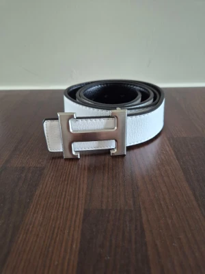H bälte - Snyggt vändbart bälte i svart och vitt med ett stort H-format metallspänne. Bältet har en stilren design och är tillverkat i skinn med struktur. Perfekt accessoar för att lyfta din outfit.