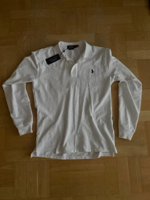 Vit långärmad piké från Polo Ralph Lauren - Klassisk vit långärmad pikétröja från Polo Ralph Lauren med broderad marinblå logga på bröstet. Skjortan har krage och knäppning vid halsen. Tillverkad i mjukt bomullsmaterial som känns skönt mot huden.