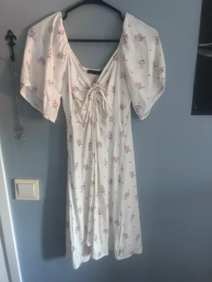 Vit blommig klänning med snörning - Söt vit klänning med rosa blommor, v-ringning och snörning framtill. Klänningen har korta, vida ärmar och ett luftigt fall. Perfekt för varma dagar och har en romantisk känsla.