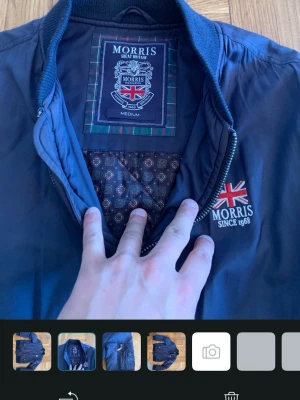 Marinblå bomberjacka från Morris - Snygg marinblå bomberjacka från Morris med Union Jack-detalj och broderad logga på bröstet. Jackan har ribbade muddar, dragkedja, två framfickor med knappar och en innerficka med mönstrat foder. Perfekt för dig som gillar klassisk brittisk stil.