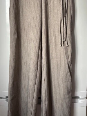 Beige linnebyxor med knytband - Snygga beige linnebyxor med diskret fiskbensmönster och smala ränder. Byxorna har vida ben och ett brett midjeband med knytband i sidan för extra stilpoäng. Perfekta för en chill och avslappnad look.