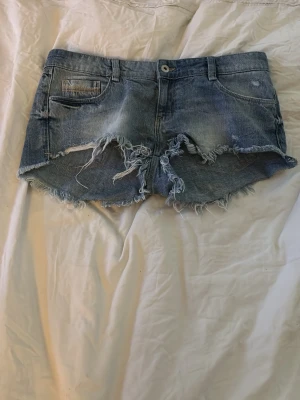 Blå slitna jeansshorts med fransar - Säljer ett par blå jeansshorts med råa, fransiga kanter och slitningar framtill. Shortsen har klassisk femficksdesign, bälteshällor och knappstängning. Perfekta för sommaren och ger en avslappnad streetstyle-look.