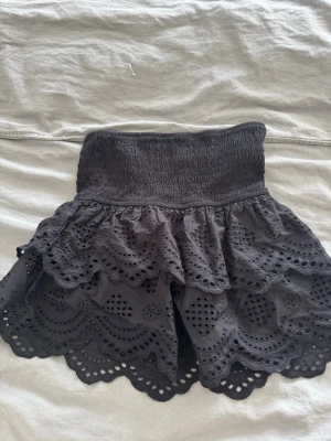Svart volangkjol från Stradivarius - Supersöt svart kortkjol från Stradivarius med bred smock i midjan och dubbla volanger. Kjolen har broderade detaljer och vågig kant nertill som ger en extra snygg look. Perfekt att styla med en enkel topp eller hoodie.