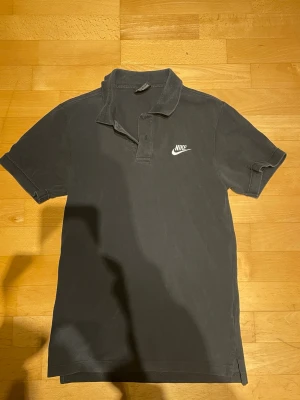 Svart pikétröja från Nike XS - Klassisk svart pikétröja från Nike med vit logga på bröstet. Tröjan har krage, korta ärmar och knäppning framtill. Perfekt för en sportig och avslappnad stil.
