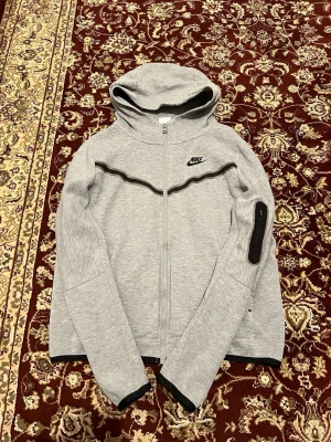 Grå Nike hoodie med dragkedja XL - Snygg grå hoodie från Nike med dragkedja framtill och svart detalj över bröstet. Huva och ficka på ärmen med dragkedja ger extra stilpoäng. Perfekt för chill dagar eller när du vill ha en sportig look. Tillverkad i mjukt material för skön känsla.